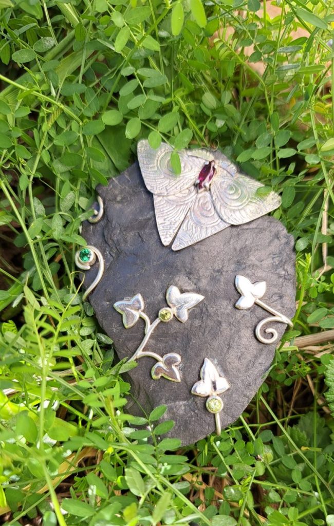 Artisans_buddies_challenge25: fertiger Anhänger aus roher Schieferplatte mit einem silbernen Falter/Schmetterling mit eingefasster Rhodolithnavette als Körper, Efeublättern und Ranken mit zwei facettierten Peridoten und einem facettierten, dunkelgrünen Topas auf der Vorderseite